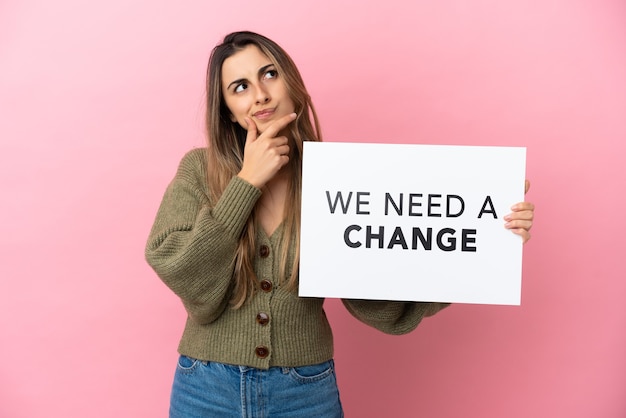 Young caucasian woman isolated on pink background holding a placard with text We Need a Change and thinking