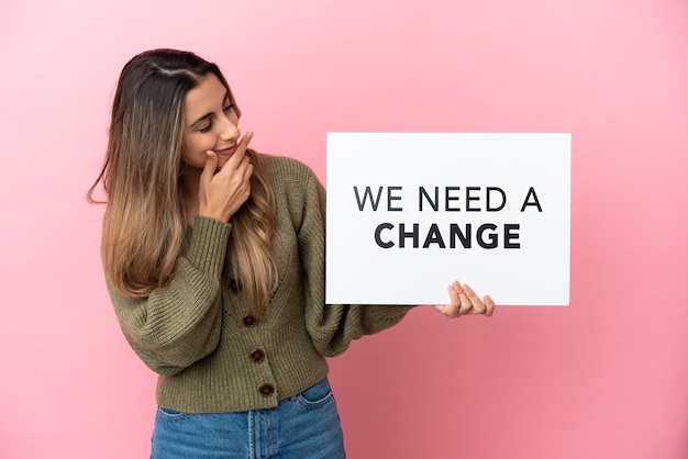 Young caucasian woman isolated on pink background holding a placard with text We Need a Change and thinking