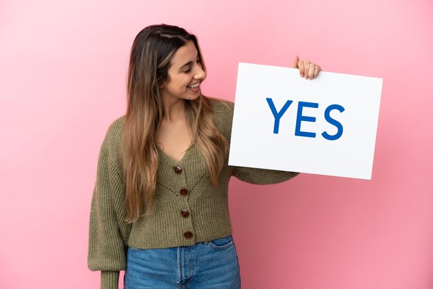 Young caucasian woman isolated on pink background holding a placard with text YES