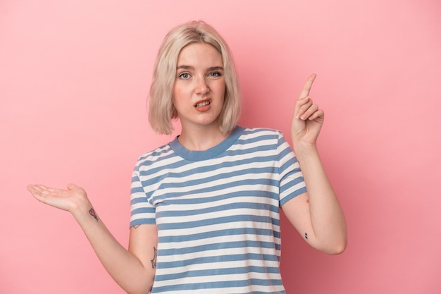 Young caucasian woman isolated on pink background holding and showing a product on hand.