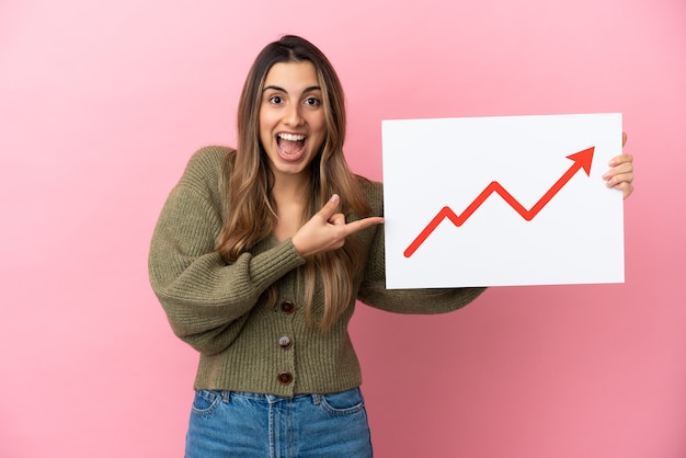 Young caucasian woman isolated on pink background holding a sign with a growing statistics arrow symbol with surprised expression