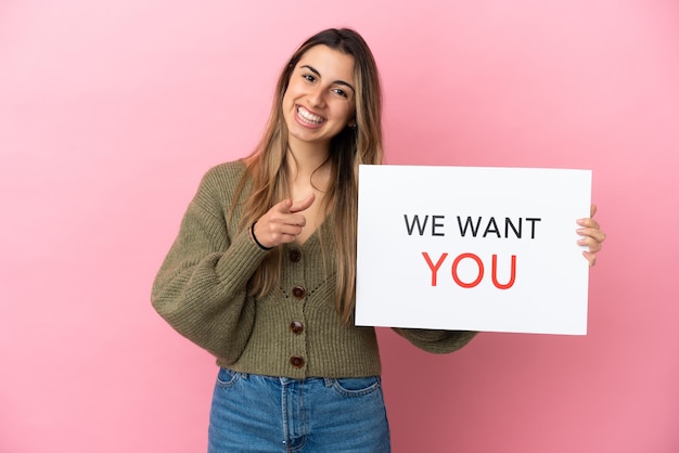 Young caucasian woman isolated on pink background holding We Want You board and pointing to the front