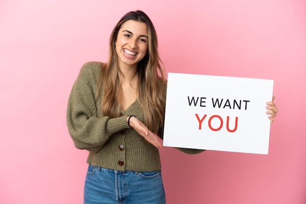 Young caucasian woman isolated on pink background holding We Want You board and  pointing it