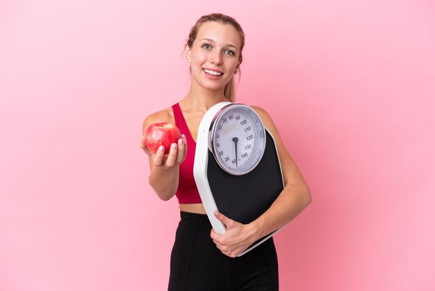 Young caucasian woman isolated on pink background holding weighing machine and offering an apple