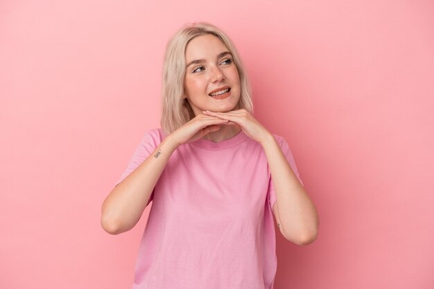 Young caucasian woman isolated on pink background keeps hands under chin, is looking happily aside.