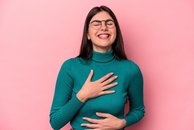 La giovane donna caucasica isolata su sfondo rosa ride felice e si diverte a tenere le mani sullo stomaco