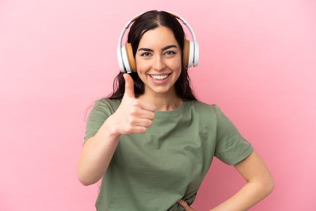 Young caucasian woman isolated on pink background listening music and with thumb up