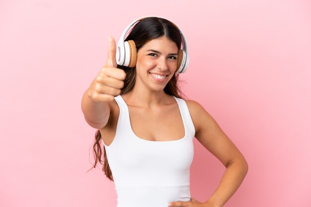 Young caucasian woman isolated on pink background listening music and with thumb up