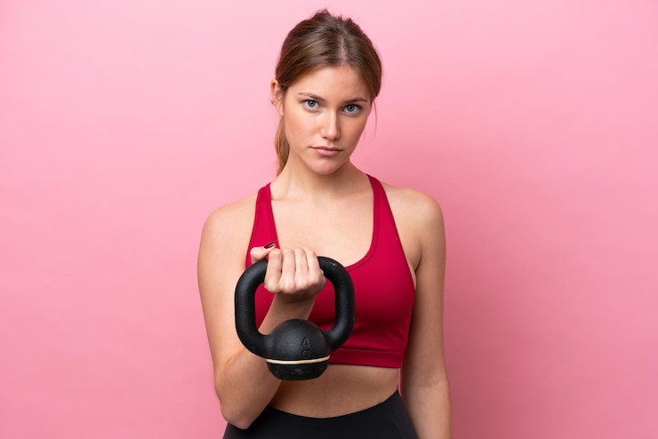 Photo young caucasian woman isolated on pink background making weightlifting with kettlebell and looking to the front