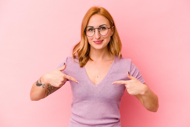 Young caucasian woman isolated on pink background person pointing by hand to a shirt copy space, proud and confident