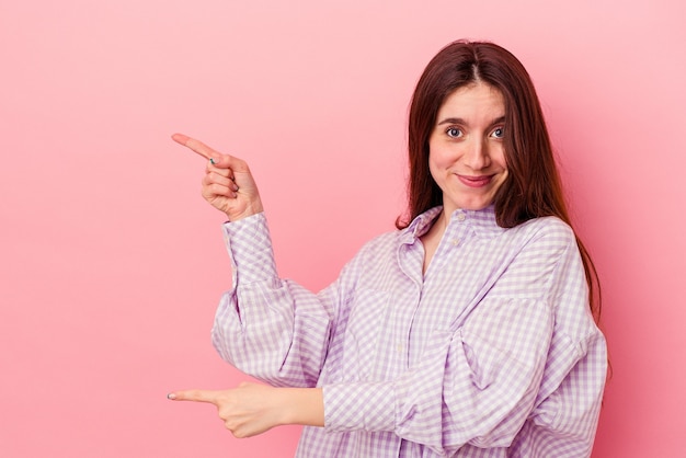 Young caucasian woman isolated on pink background pointing with forefingers to a copy space, expressing excitement and desire.