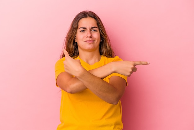 Young caucasian woman isolated on pink background  points sideways, is trying to choose between two options.