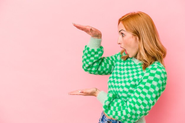 Foto giovane donna caucasica isolata su sfondo rosa scioccata e stupita in possesso di uno spazio di copia tra le mani.