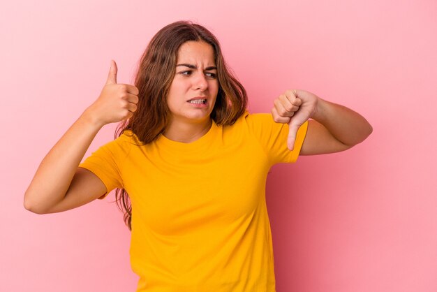 Young caucasian woman isolated on pink background  showing thumbs up and thumbs down, difficult choose concept