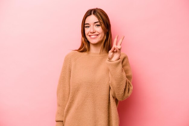 Young caucasian woman isolated on pink background showing victory sign and smiling broadly