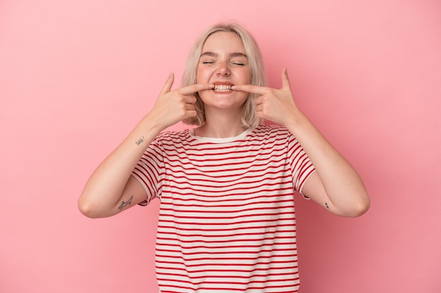 Young caucasian woman isolated on pink background smiles, pointing fingers at mouth.