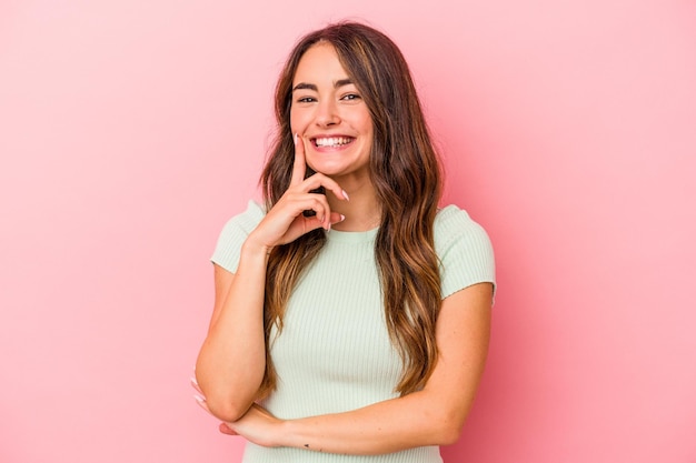 Giovane donna caucasica isolata su sfondo rosa sorridente felice e sicura di sé, toccando il mento con la mano.