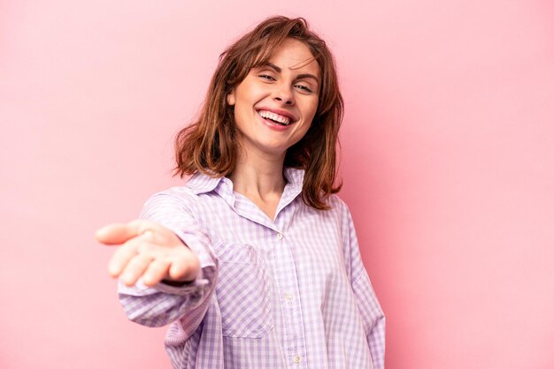 Young caucasian woman isolated on pink background stretching hand at camera in greeting gesture