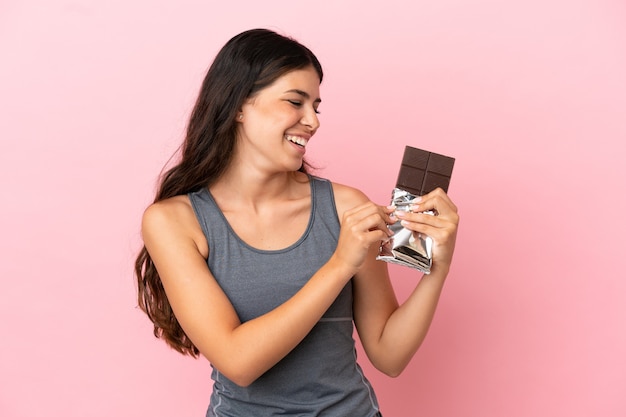 Young caucasian woman isolated on pink background taking a chocolate tablet and happy