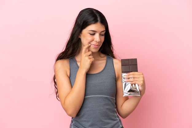 Young caucasian woman isolated on pink background taking a chocolate tablet and having doubts