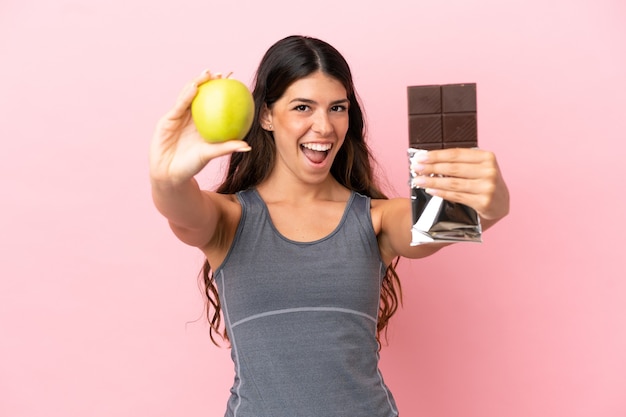 Young caucasian woman isolated on pink background taking a chocolate tablet in one hand and an apple in the other