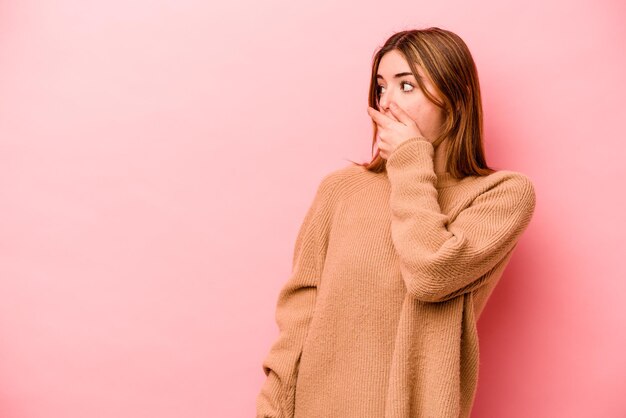 Young caucasian woman isolated on pink background thoughtful looking to a copy space covering mouth with hand