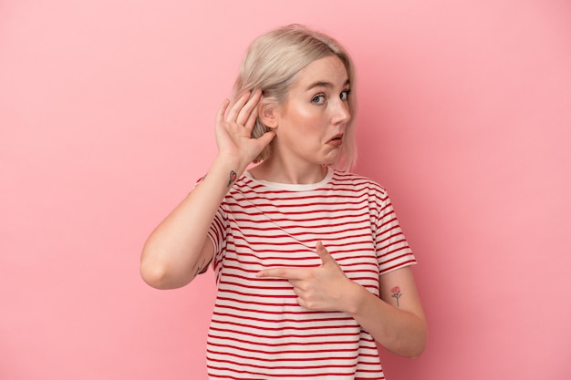 Young caucasian woman isolated on pink background trying to listening a gossip.