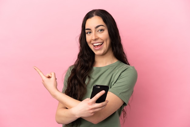 Young caucasian woman isolated on pink background using mobile phone and pointing back