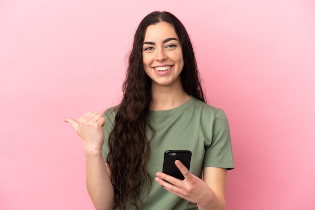 Young caucasian woman isolated on pink background using mobile phone and pointing to the lateral