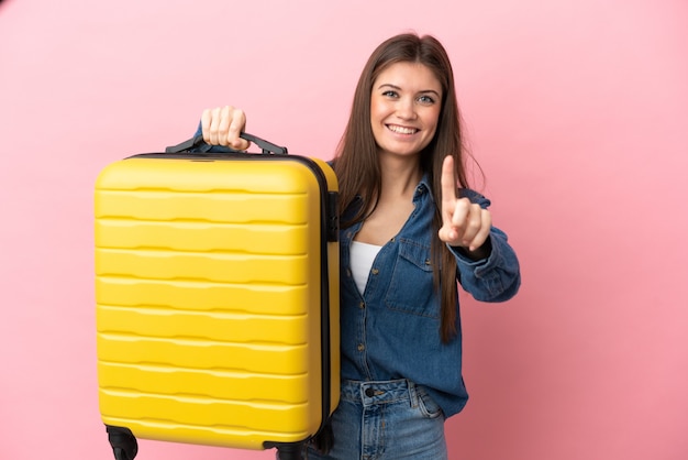 Giovane donna caucasica isolata su sfondo rosa in vacanza con valigia da viaggio e contando uno