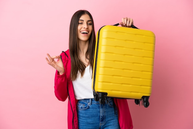 Young caucasian woman isolated on pink background in vacation with travel suitcase and making OK sign