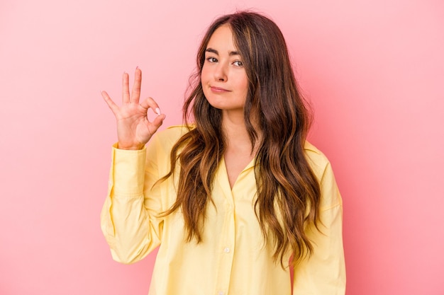 Young caucasian woman isolated on pink background winks an eye and holds an okay gesture with hand.