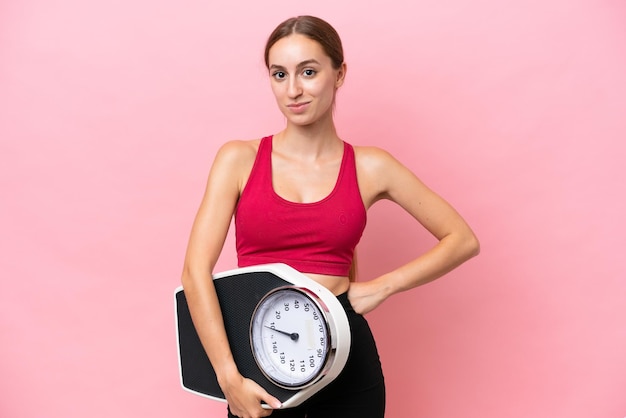 Young caucasian woman isolated on pink background with arms at hip and holding weighing machine