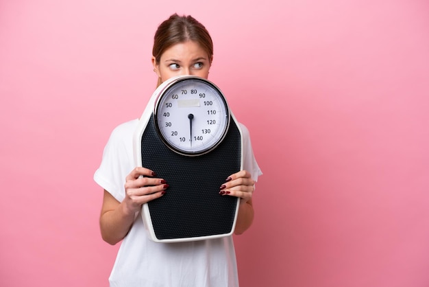 Young caucasian woman isolated on pink background with weighing machine and hiding behind it