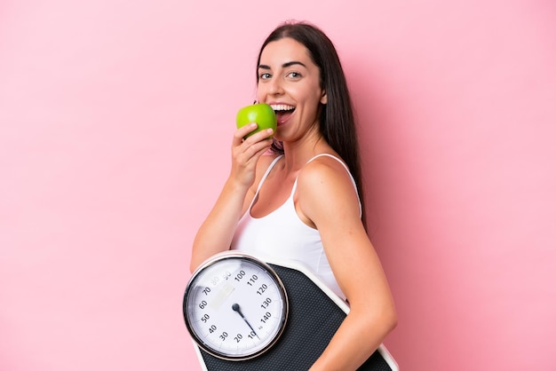 Young caucasian woman isolated on pink background with weighing machine and with an apple