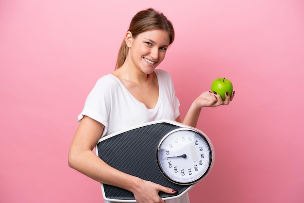 Young caucasian woman isolated on pink background with weighing machine and with an apple