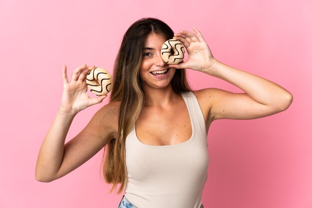 Young caucasian woman isolated on pink wall holding a donut in an eye