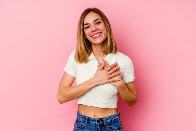 Giovane donna caucasica isolata sulla parete rosa che ride mantenendo le mani sul cuore