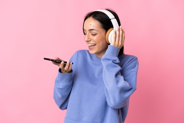 Young caucasian woman isolated on pink wall listening music with a mobile and singing