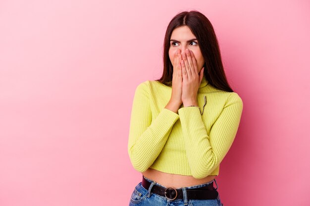 Young caucasian woman isolated on pink wall thoughtful looking to a copy space covering mouth with hand.