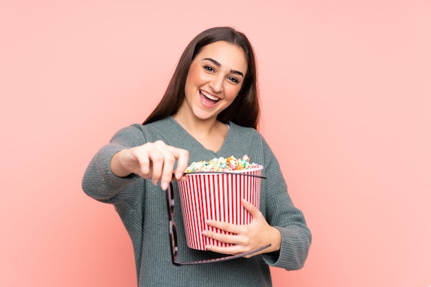 Young caucasian woman isolated on pink with 3d glasses and holding a big bucket of popcorns
