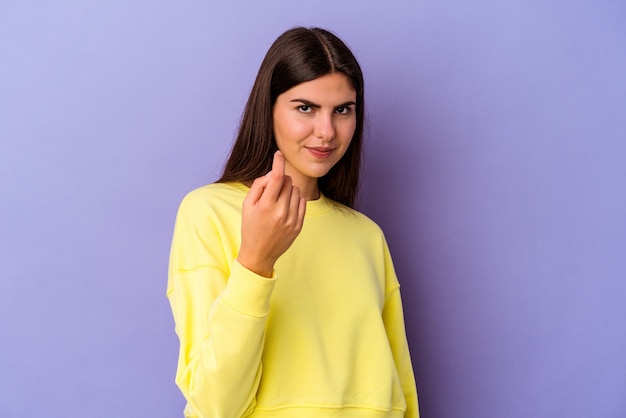 Young caucasian woman isolated on purple background pointing with finger at you as if inviting come closer.