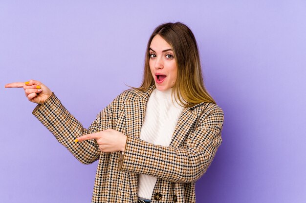 Young caucasian woman isolated on purple background pointing with forefingers to a copy space, expressing excitement and desire.