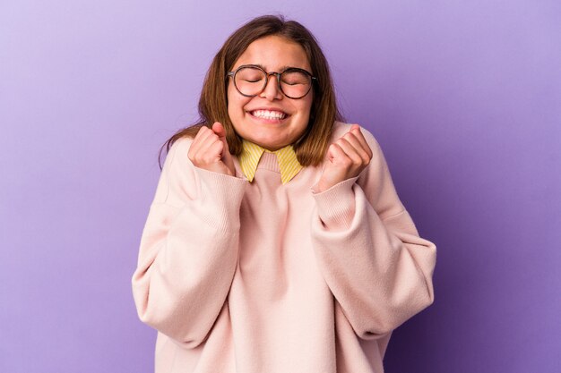 Giovane donna caucasica isolata su sfondo viola alzando il pugno, sentendosi felice e di successo. concetto di vittoria.