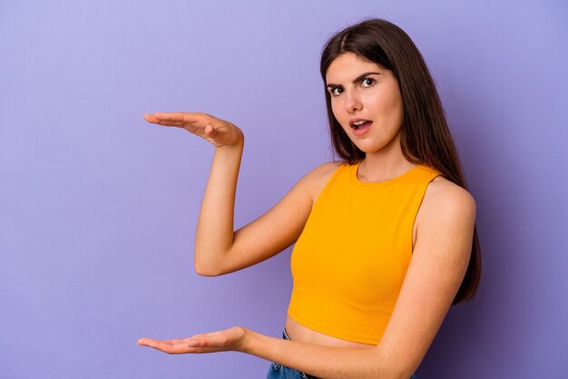 Young caucasian woman isolated on purple background shocked and amazed holding a copy space between hands.