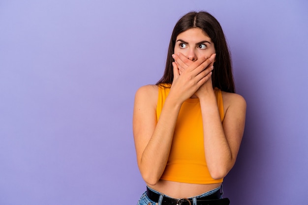 Young caucasian woman isolated on purple background thoughtful looking to a copy space covering mouth with hand.