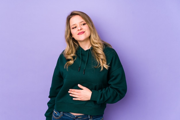 Young caucasian woman isolated on purple background touches tummy, smiles gently, eating and satisfaction concept.