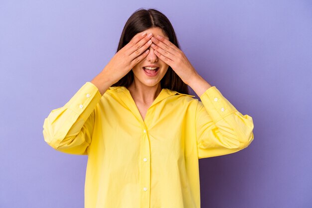 Young caucasian woman isolated on purple wall covers eyes with hands, smiles broadly waiting for a surprise.