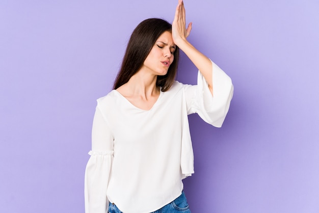 Young caucasian woman isolated on purple wall forgetting something, slapping forehead with palm and closing eyes.