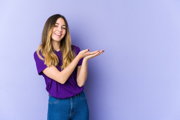 Young caucasian woman isolated on purple wall holding a copy space on a palm.